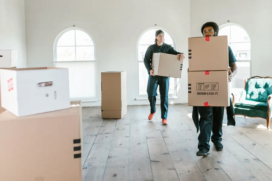 Two movers carrying cardboard moving boxes across an empty bright room with arched windows and wood floor