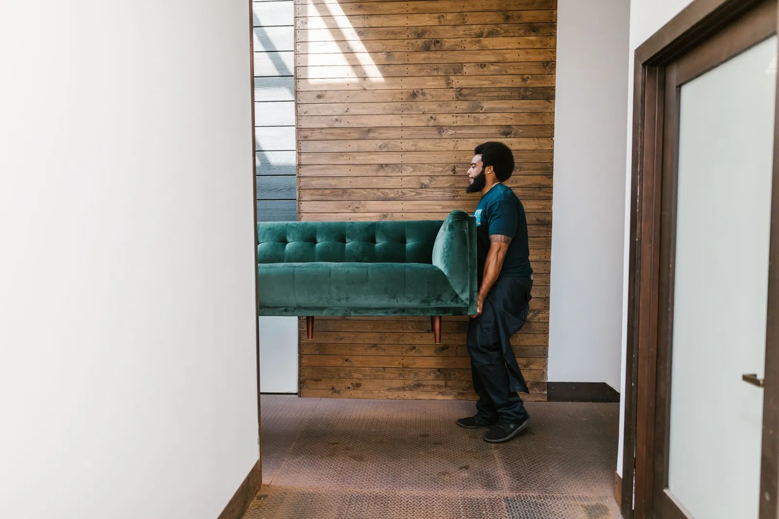 Single Black mover carrying a green velvet sofa through a hallway with a warm wood-slat accent wall and a white door
