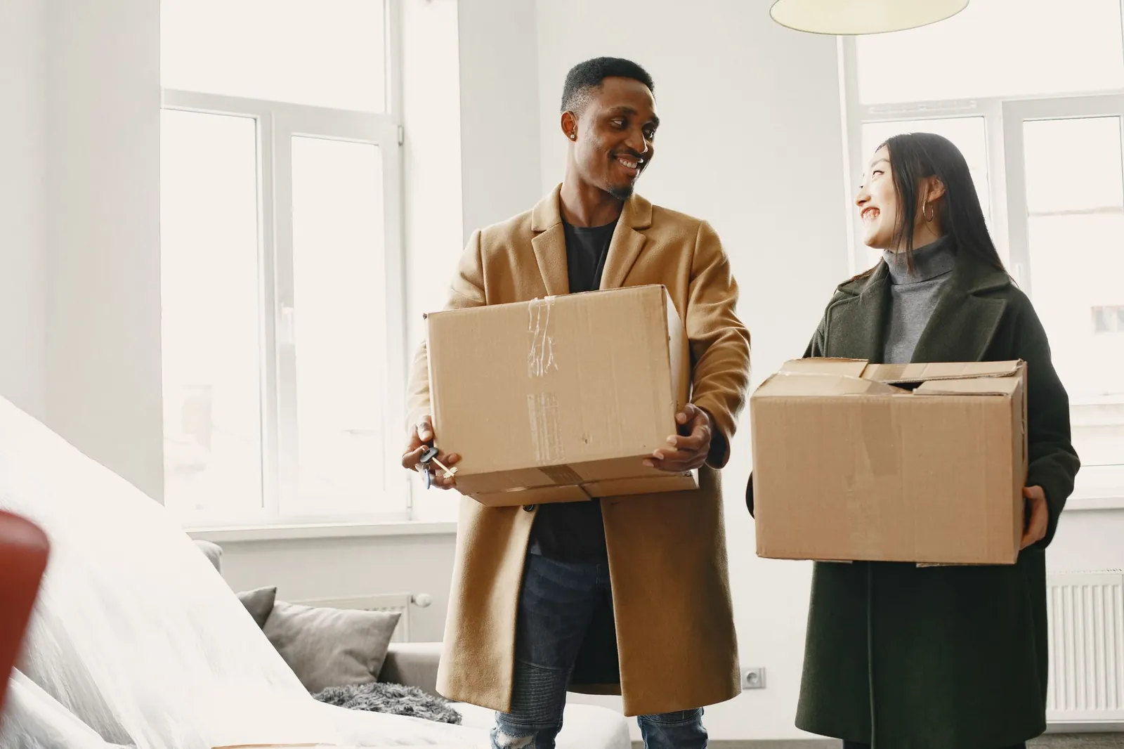 Black man in camel overcoat and Asian woman in dark green coat smiling at each other while holding moving boxes in a bright white apartment 