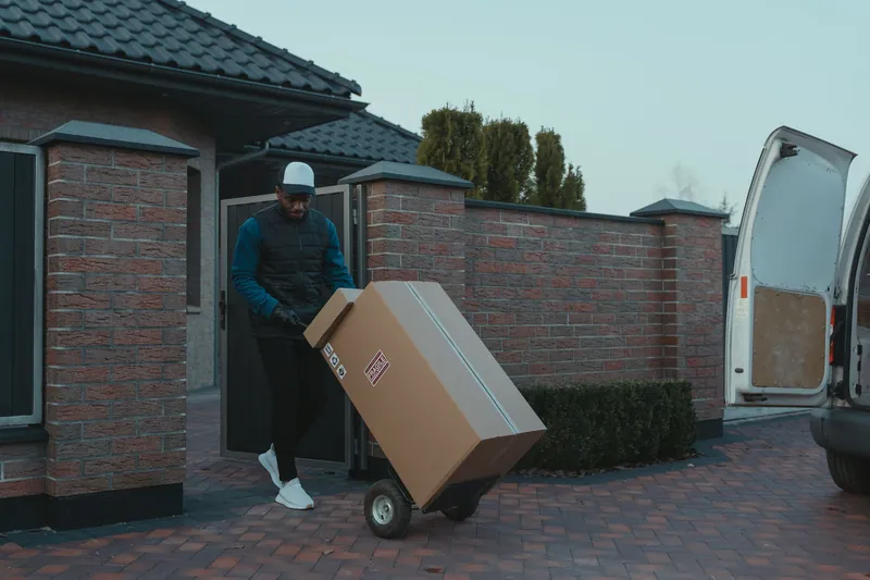 Black delivery/moving worker wheeling a large 'FRAGILE' box on a hand truck through a brick-paved driveway beside a white van with open rear