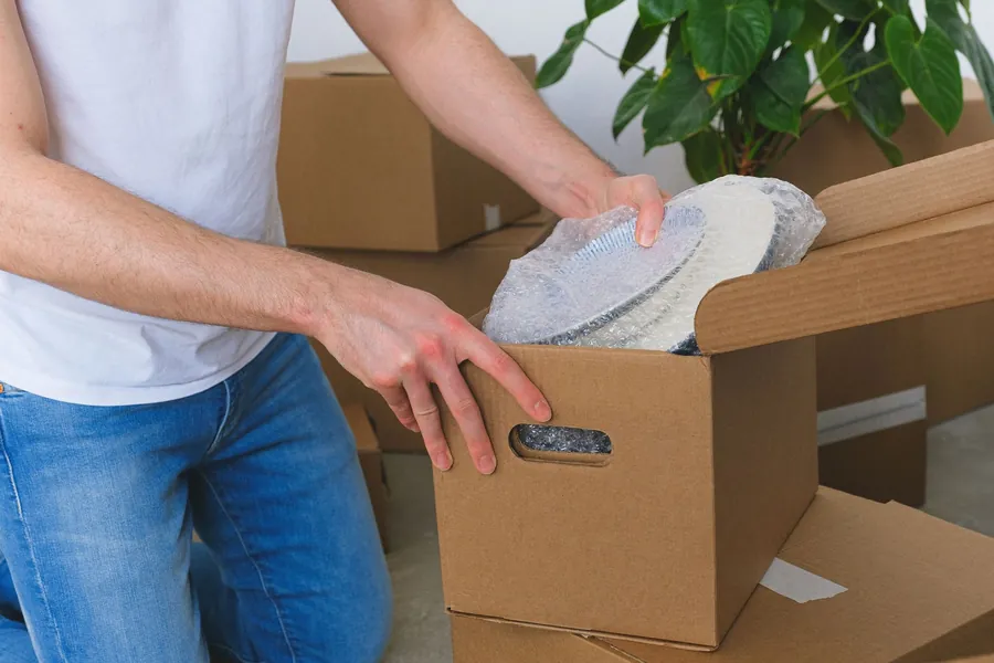 Close crop of a man in white t-shirt and blue jeans placing bubble-wrapped plates into a cardboard box with handle cut-outs