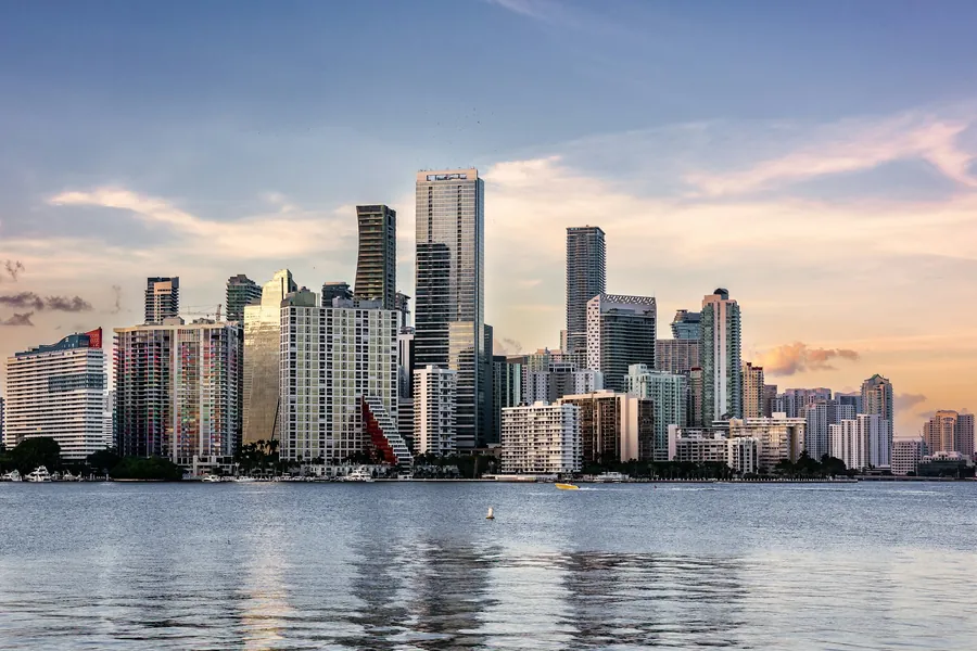 Brickell / downtown Miami skyline across Biscayne Bay at golden hour, pastel pink-orange clouds, calm water reflections