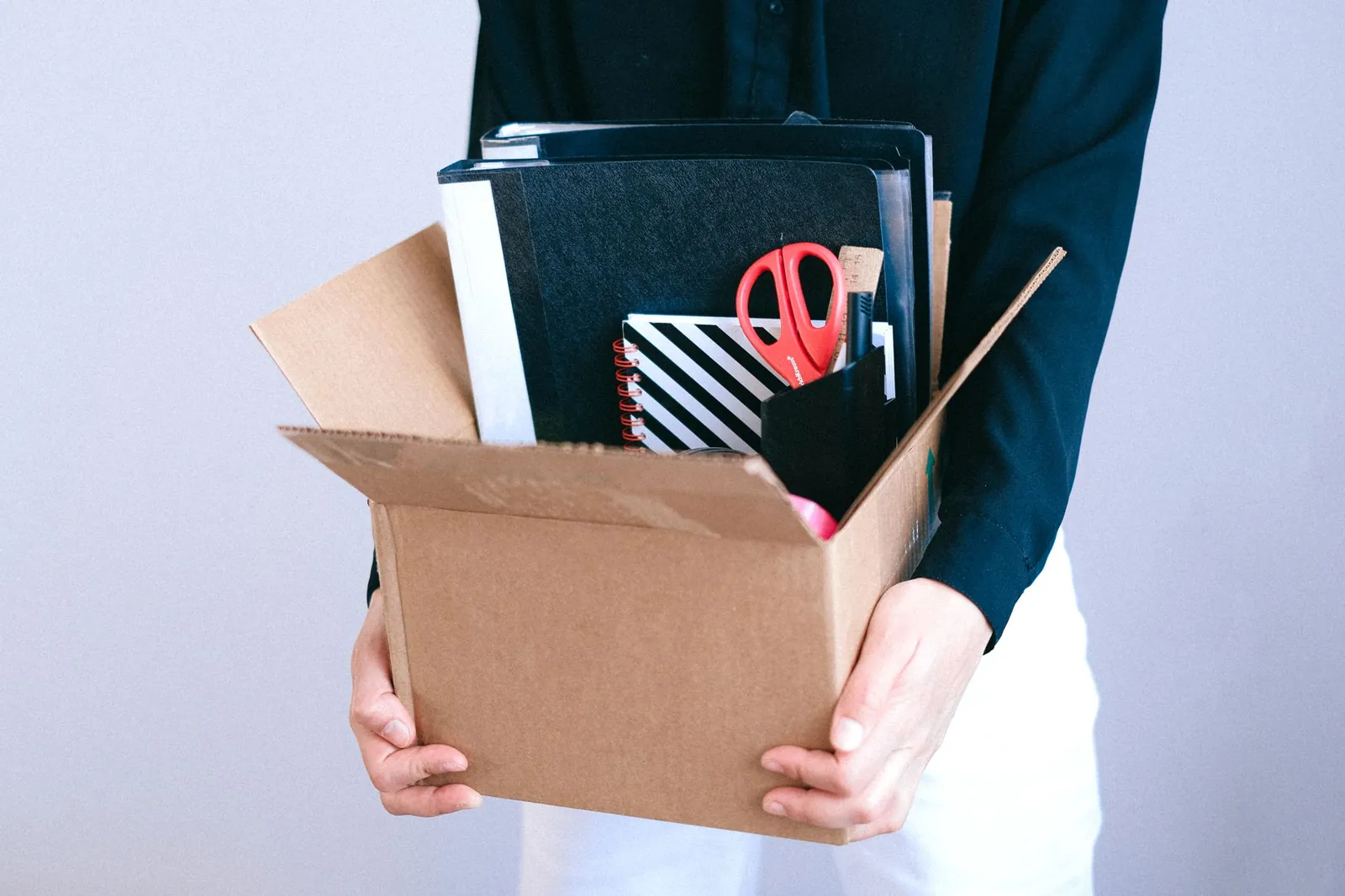 Torso crop of a person in a black long-sleeve top and white pants holding a cardboard box of office supplies (binders, notebooks, red scisso