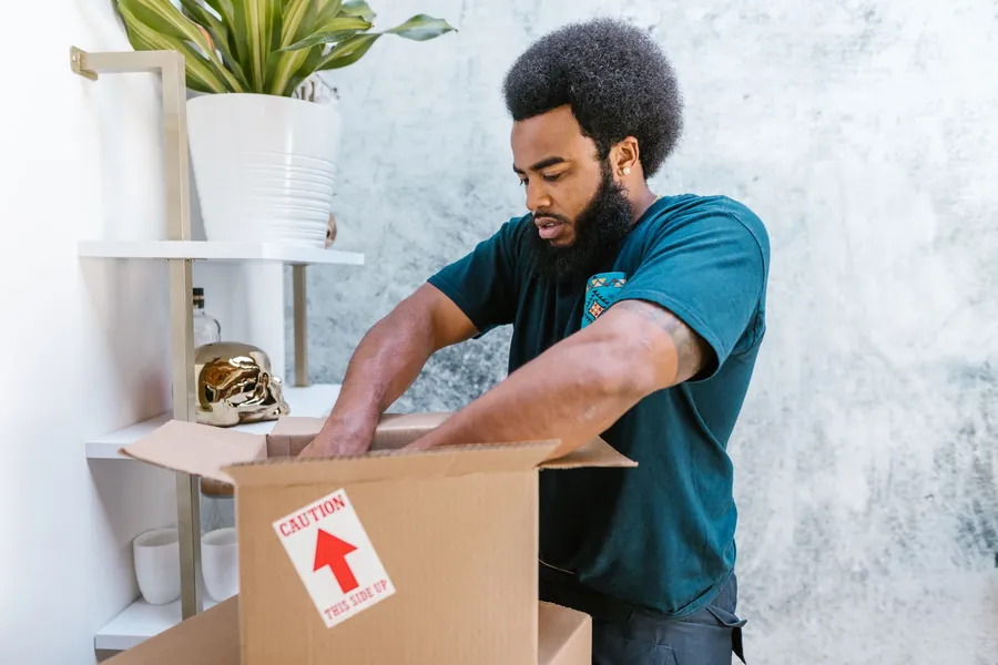 Black mover with afro and beard packing items into a cardboard box with a red 'CAUTION - THIS SIDE UP' arrow sticker