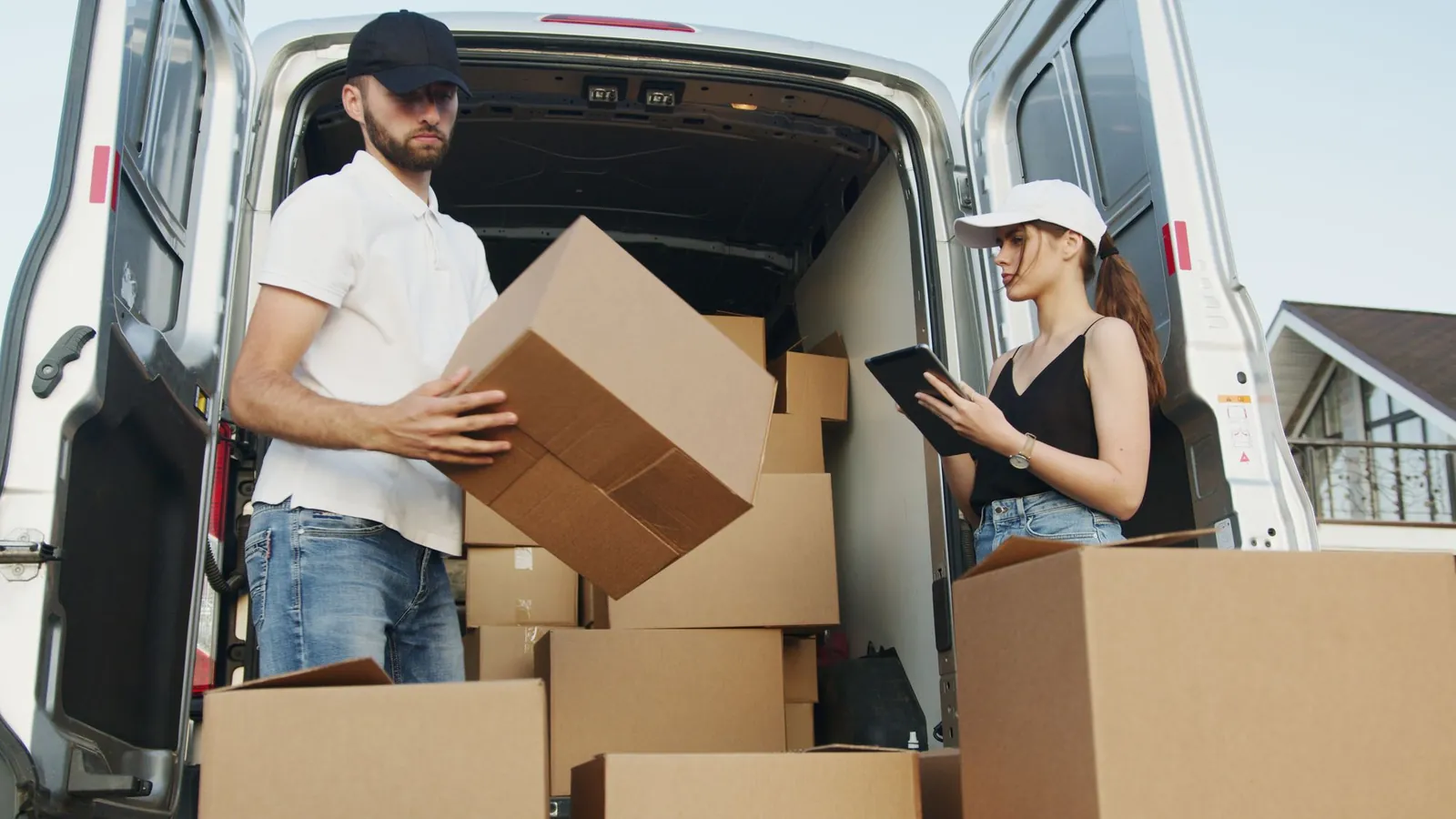 Man in white polo, jeans and black cap lifting a box at the back of a white van packed with cardboard boxes