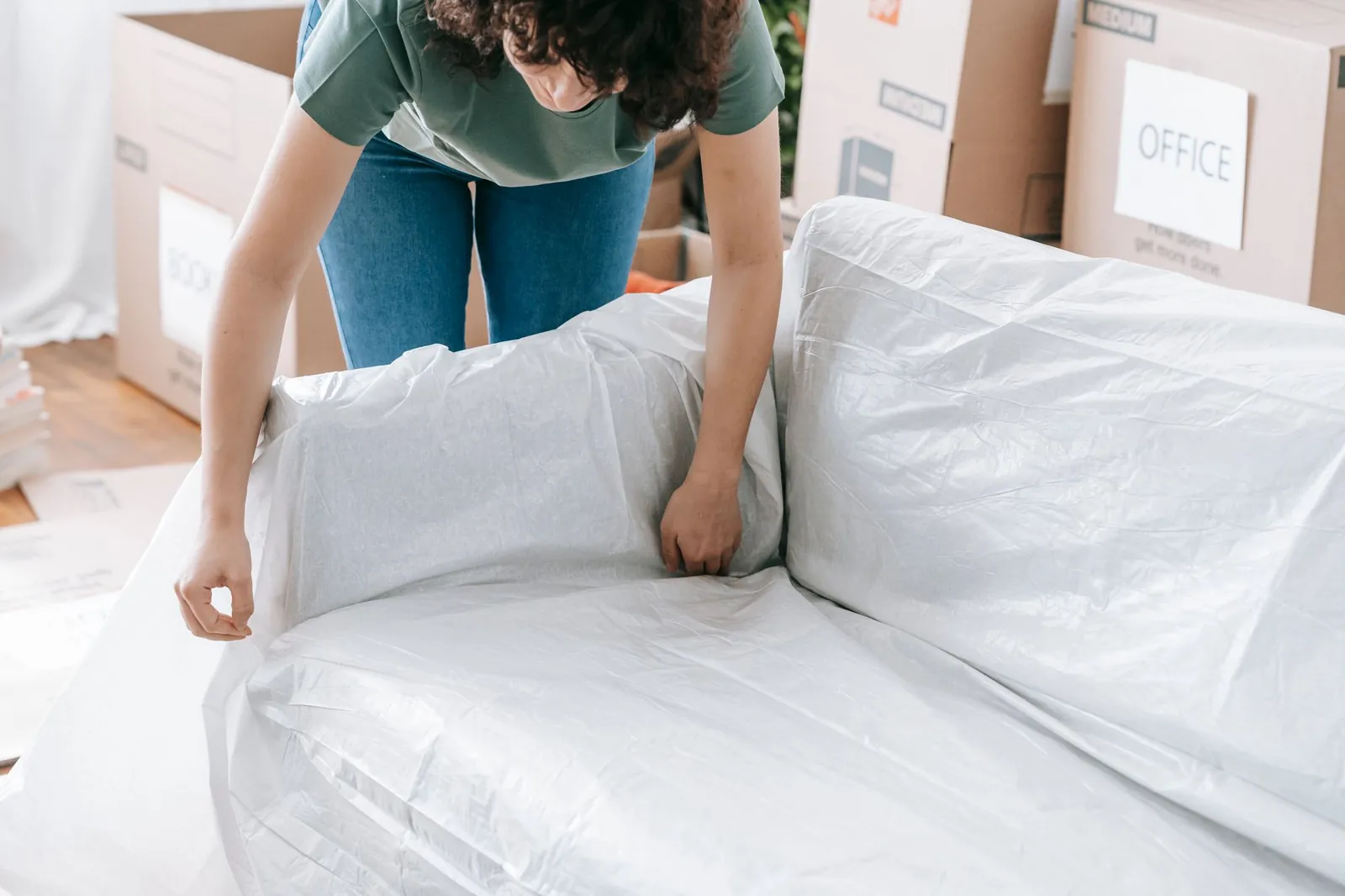 Woman with curly dark hair (sage-green t-shirt, blue jeans) tucking clear plastic stretch sheeting over a sofa